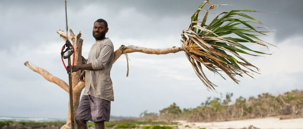 Efate, Vanuatu: Daniel stand on a beach holding a fishing spear and leaning on a withered palm tree.