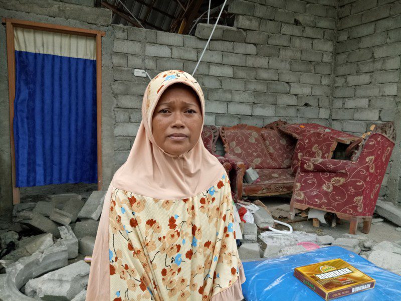 An Indonesia earthquake survivor stands outside her collapsed home in the city of Palu. She has received an emergency shelter kit from Oxfam.