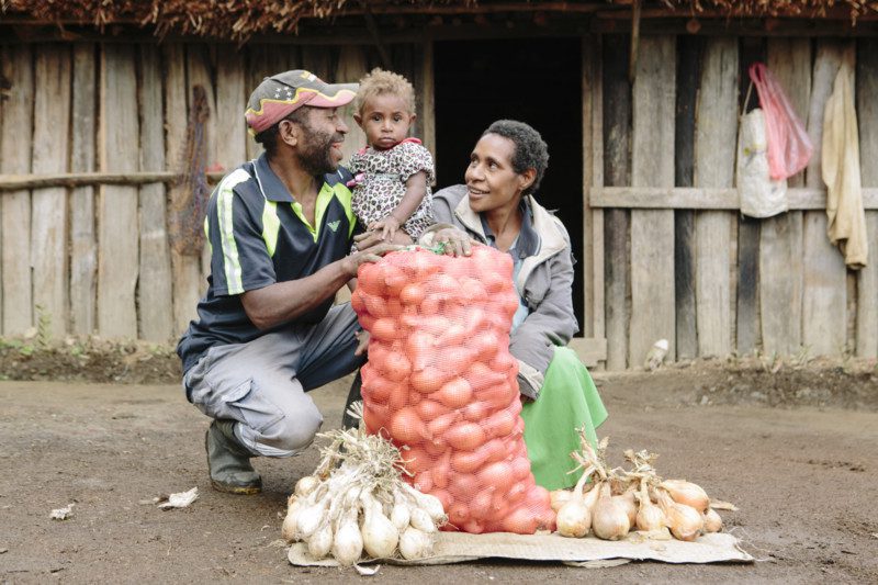 Man and woman hold child, standing behind a bag of onions smiling 
