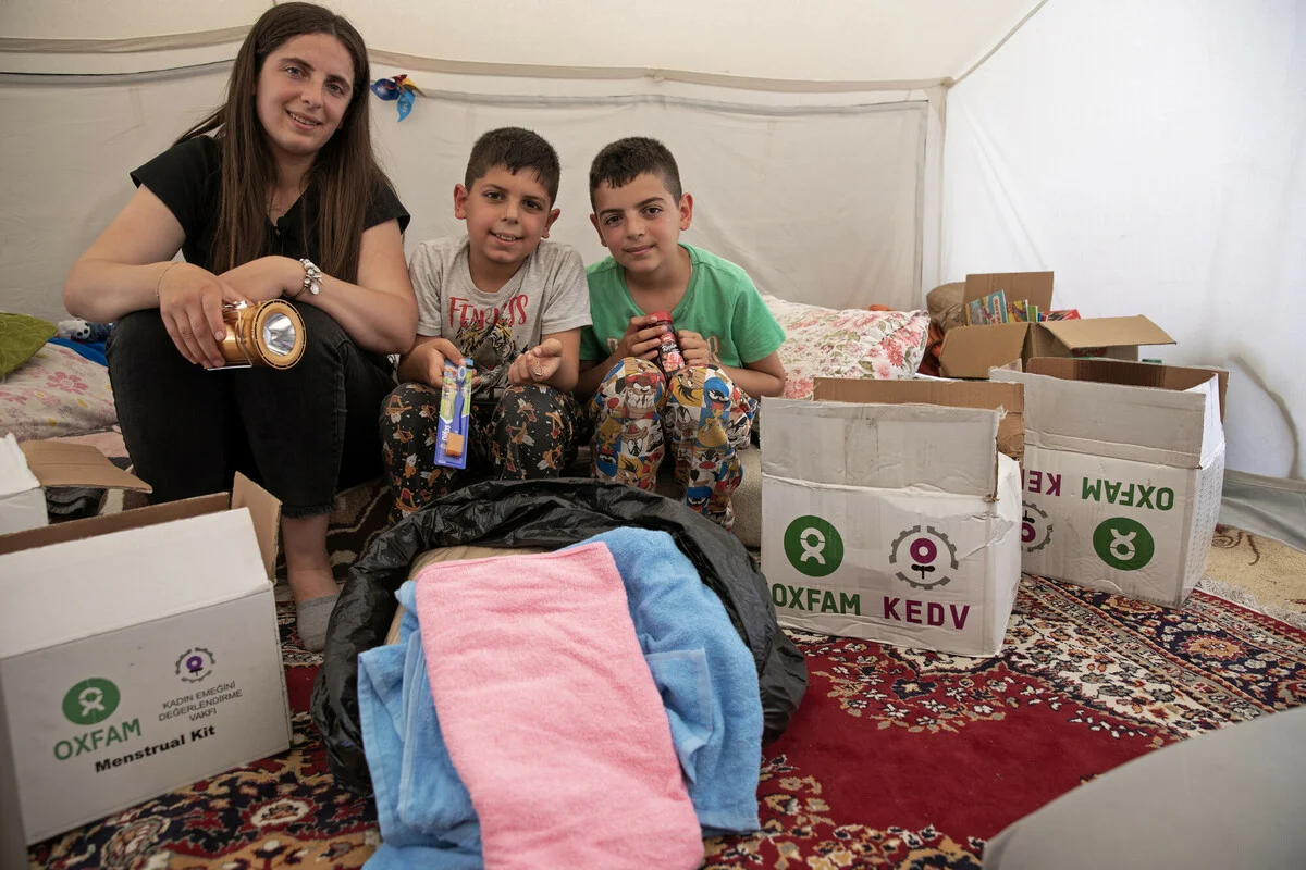 Turkiye: Mother and her two young sons are sitting in their tent. Surrounded by boxes of materials provided by Oxfam and KEDV. They are smiling and seem happy and grateful to have made it to a safe area. The little family is hugging and playing around while unboxing the supplies and goods boxes. Photo: Delizia Flaccavento/Oxfam